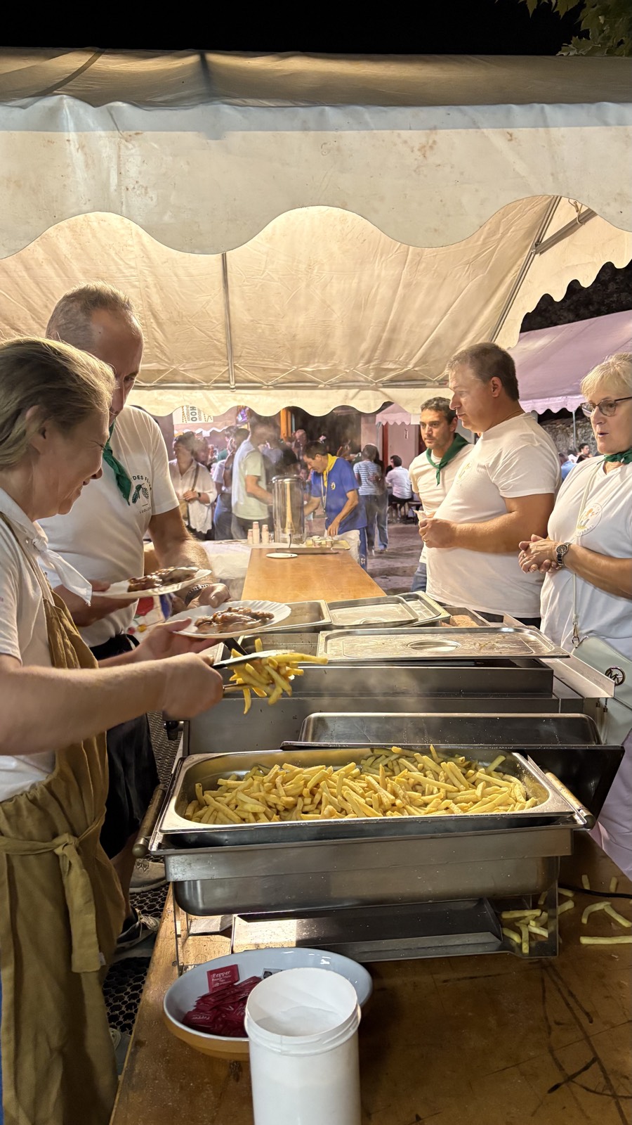 Des bénévoles servent frites et grillades derrière un buffet, sous une tente